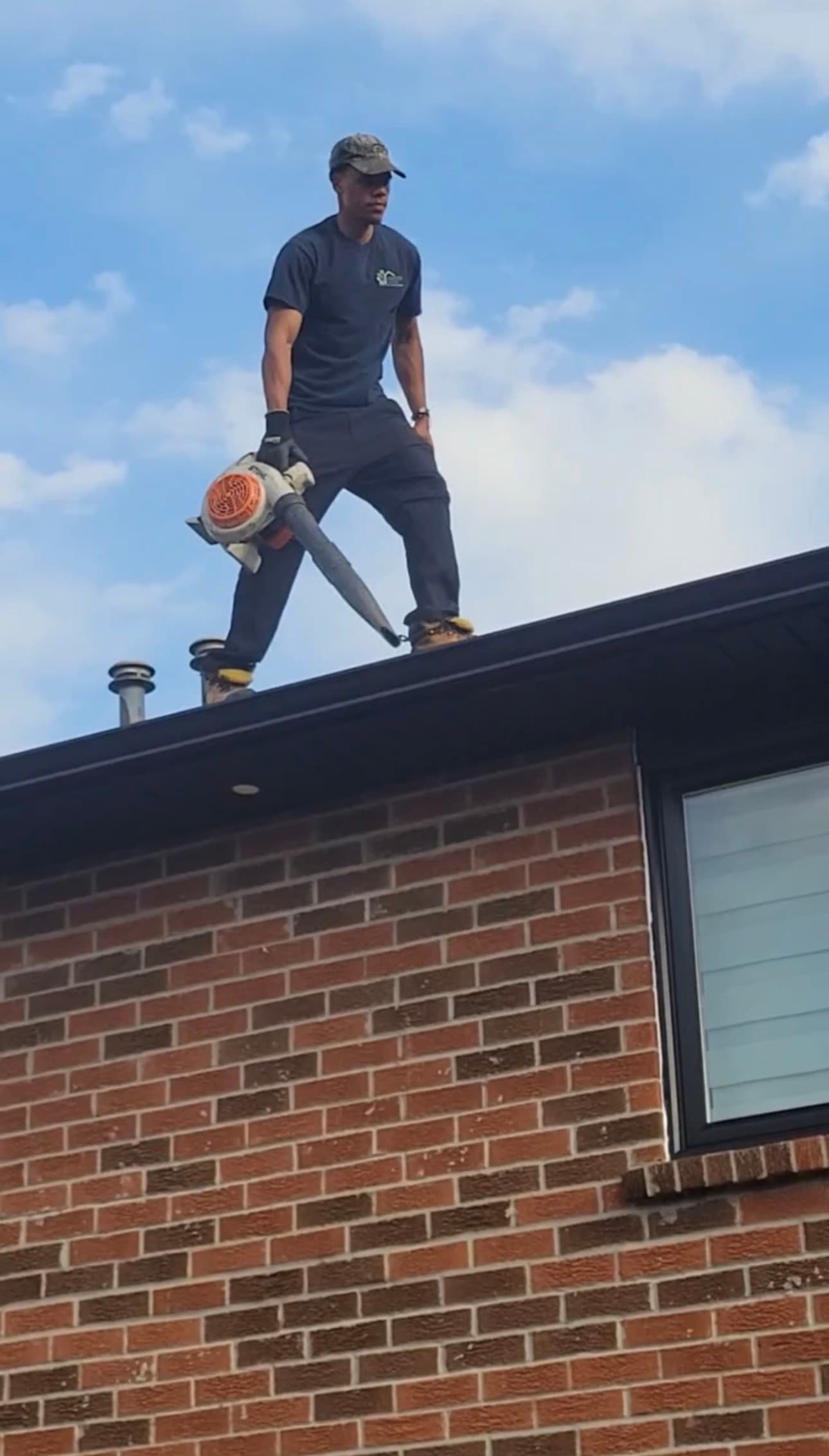 Man in work clothes standing on a brick house roof holding a leaf blower.