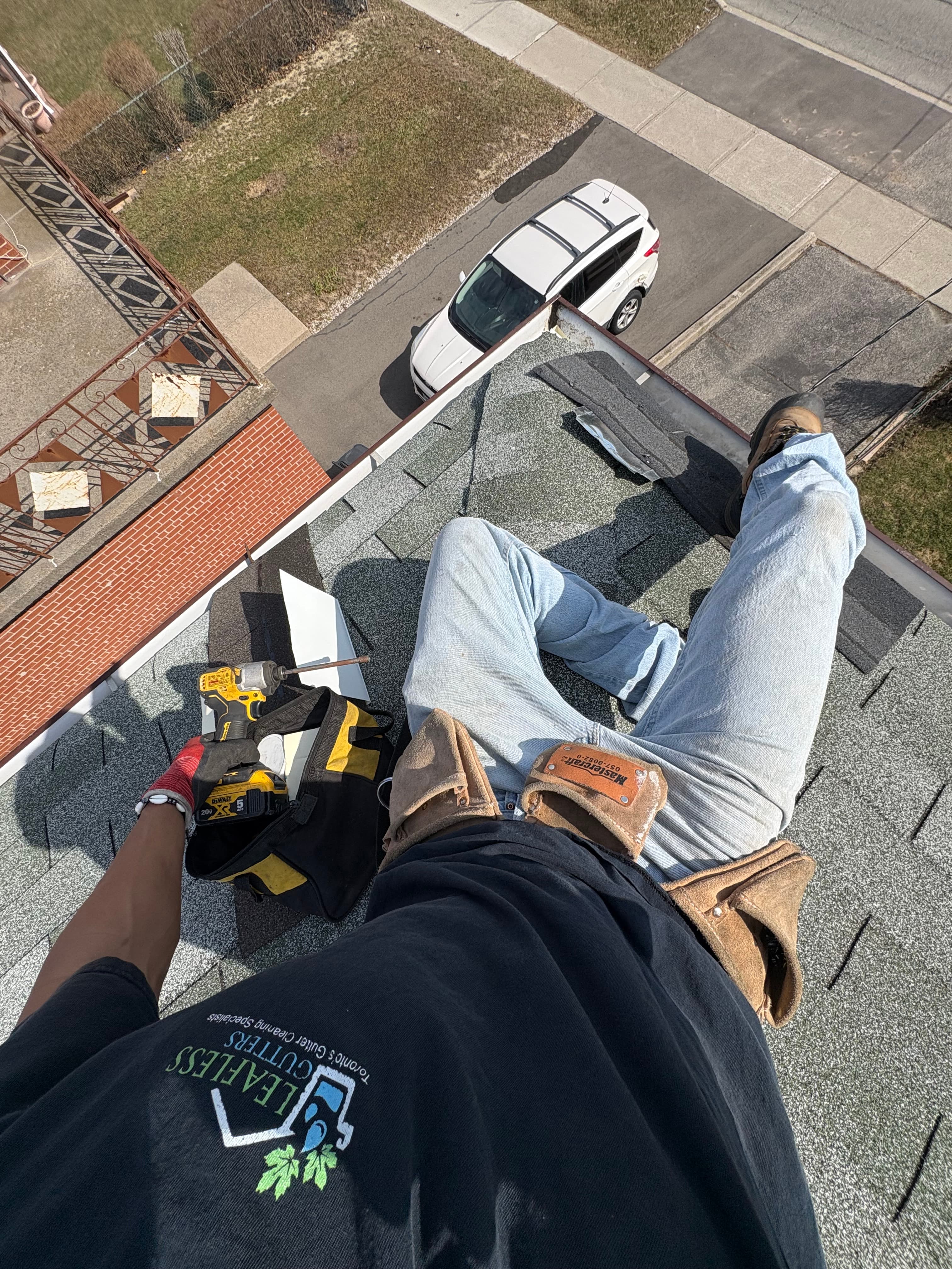 Roofer sitting on a shingled roof with tools, looking down at a white SUV.